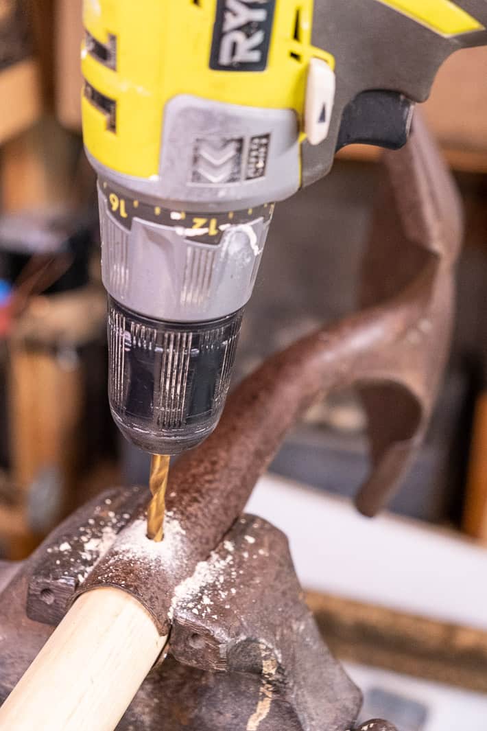 Shovel head held tightly in a vice while a drill kicks up sawdust, creating a hole in the new handle for a rivet to go through.
