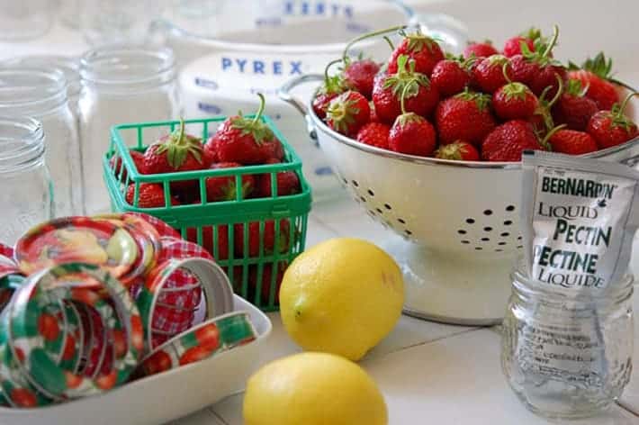 Strawberries in a white colander, lemons, pectin and other strawberry jam ingredients on a white kitchen counter.