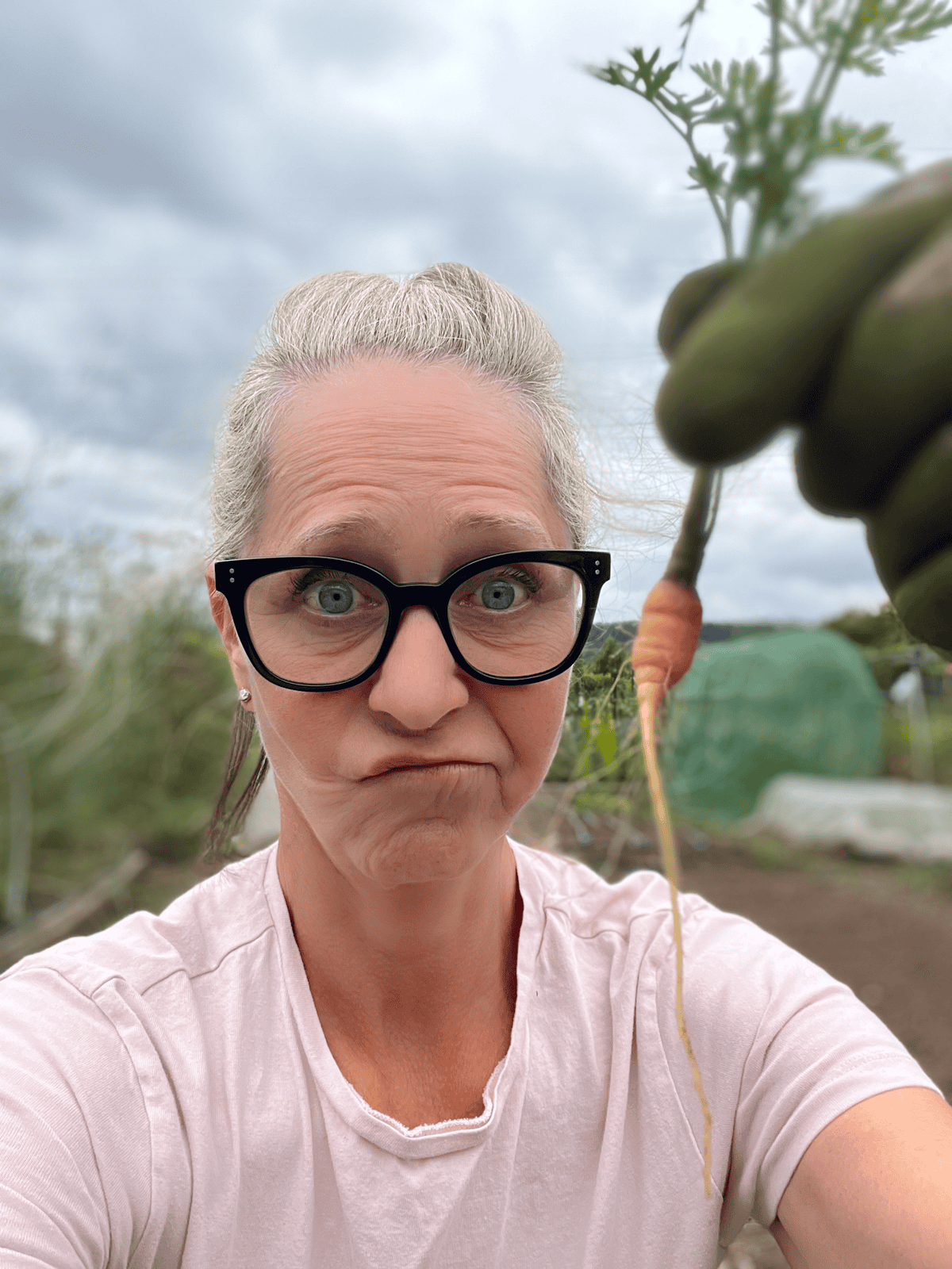 Karen Bertelsen in pink shirt and black glasses holding up a puny carrot in her garden.