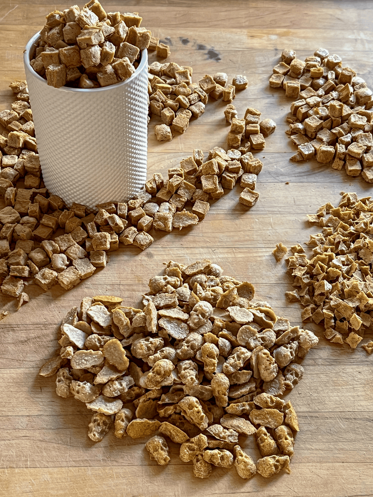 Cups and cups of dog treats on a wood butcher block countertop, spilling out of a white ceramic container. 