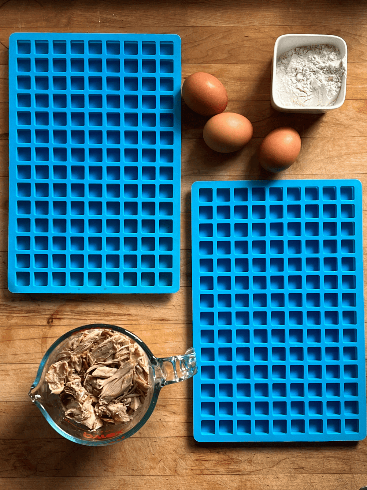 2 blue silicone treat molds, 2 cups of cooked chicken, eggs and flour on a butcher block countertop. 