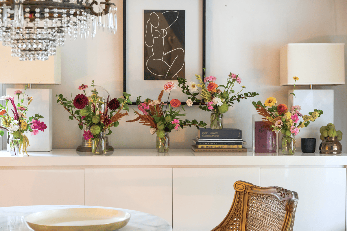 A series of 6 colourful flower arrangements in glass mason jars on a white lacquer buffet.