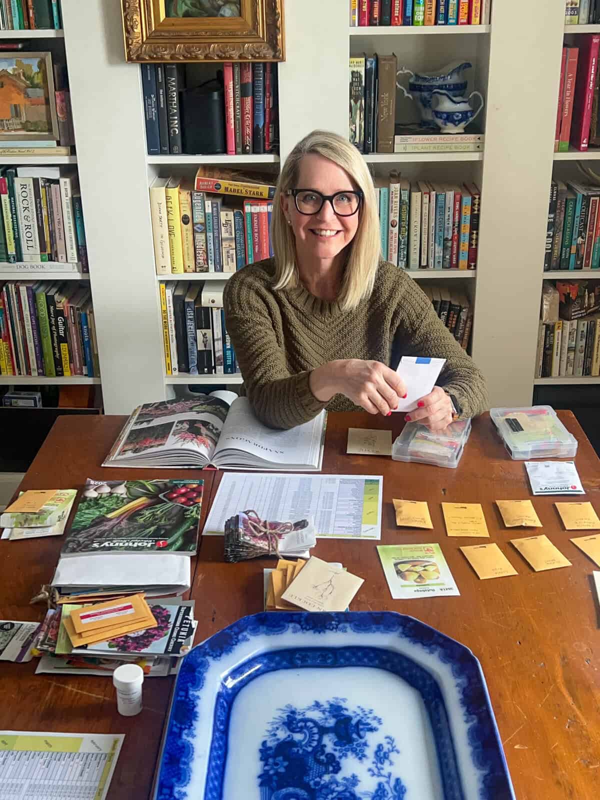 Karen Bertelsen sits at a harvest table in front of a bookcase while sorting seeds.